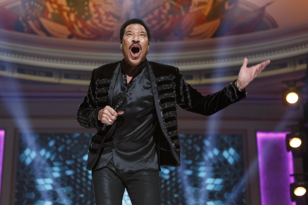Lionel Richie performs at the Grammys, wearing a black dress and holding a microphone on a brightly lit stage with a painting visible in the background.