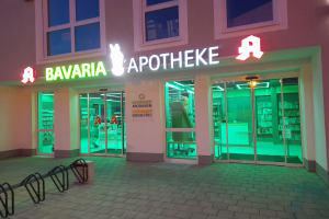 Außenansicht einer Apotheke namens "Bavaria Apotheke" mit Glastüren und -fenstern, die beleuchtete Produkte im Inneren und verstreute Gegenstände links davon zeigen.