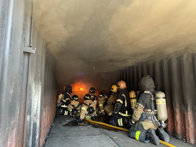 Feuerwehrleute in Helmen und Sauerstoffflaschen arbeiten in einem raucherfüllten Tunnel, einer hält ein Rohr, mit sichtbarem Feuer und einer Wand links daneben.
