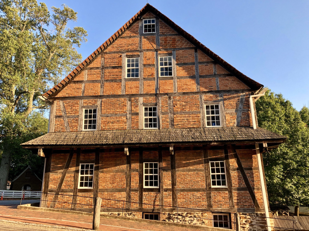 Altes Backsteingebäude mit einem zentralen Uhrenturm, umgeben von einem Zaun und einer Straße, vor einem Wald und einem klaren blauen Himmel.