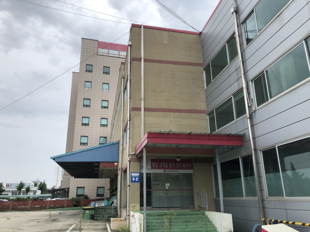 Großes modernes Gebäude mit vielen Fenstern, als Hauptsitz der Koreanischen Nationalen Wissenschaftlichen Technologie-Universität bezeichnet, umgeben von Grünflächen, einer Treppe, einem Schild, Pfosten, einem Zaun, Bäumen, Fahrzeugen, Stromleitungen und einem bewölkten Himmel.