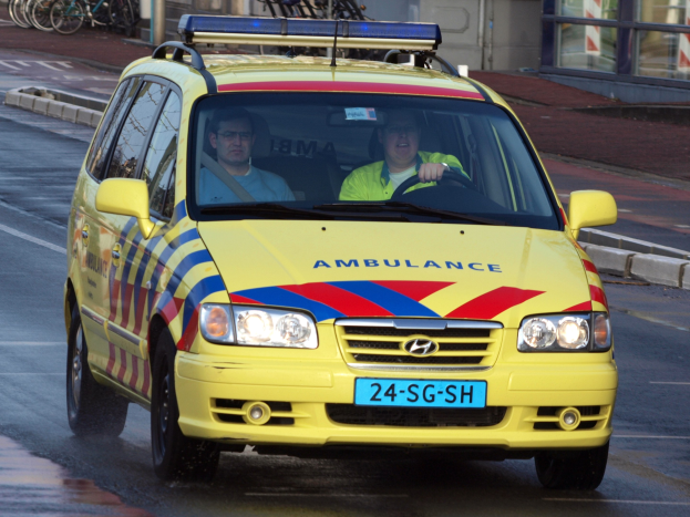 Ein gelber Rettungswagen mit zwei sichtbaren Insassen fährt an parkenden Fahrrädern auf einem Gehweg vorbei, mit einem Glasfenstergebäude im Hintergrund.