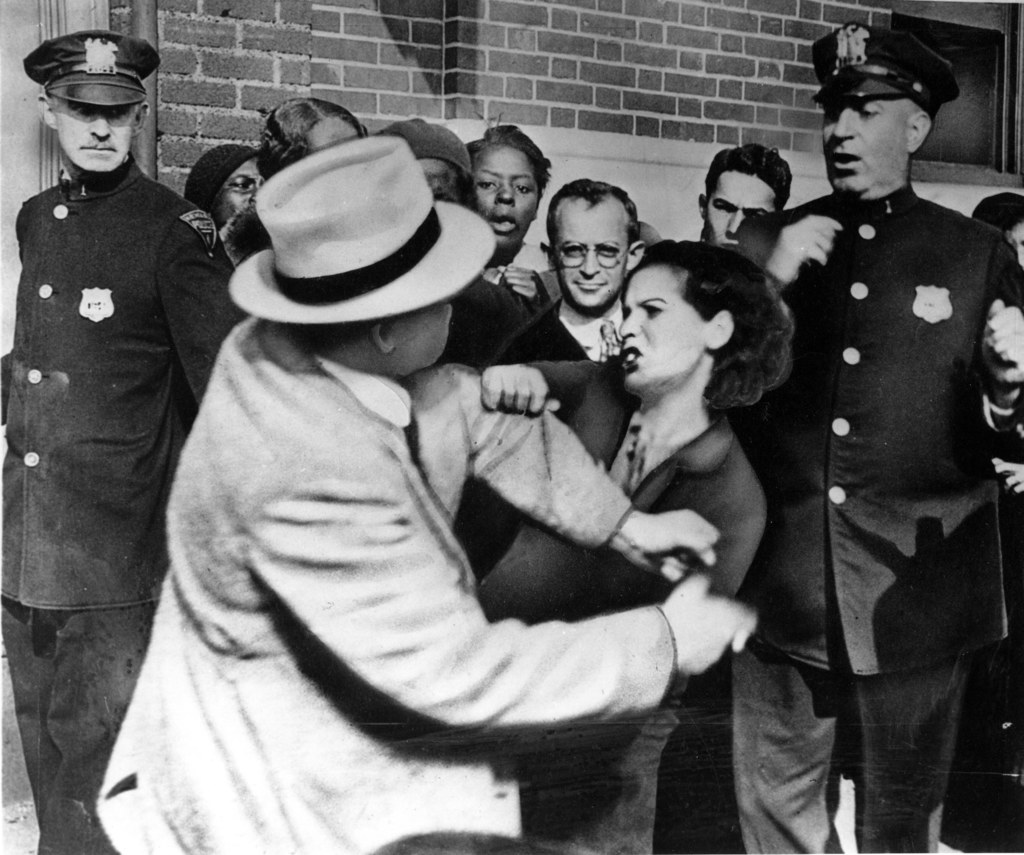 Zwei Personen kämpfen in einem Schwarz-Weiß-Bild, mit mehreren anderen im Hintergrund, einige tragen Mützen, und eine Wand dahinter.