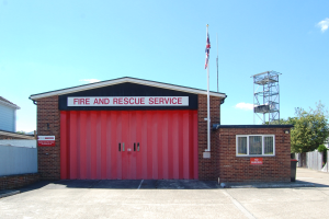 Feuerwehr- und Rettungsdienstgebäude mit roter Tür, Fenstern, einem Namensschild, einem Flaggenmast mit Flagge, einem Metallturm, einem Zaun, einer Gruppe von Bäumen und einem bewölkten Himmel.
