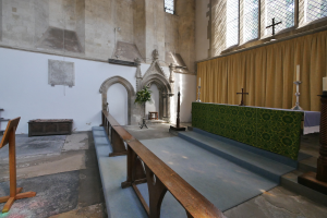 Das Innere einer Kirche mit hölzernen Kirchenbänken, einem Geländer, einem Teppichboden, einem Tisch mit Kerzen und einer Blumenvase, einem Kreuzsymbol, Vorhängen, Fenstern und gerahmten Wänden.
