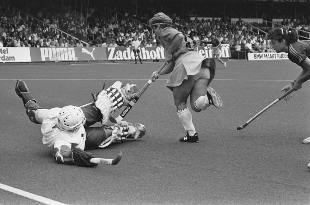 Schwarzes Bild einer Gruppe von Frauen, die auf einem Stadion Rasenfeldhockey spielen, mit Zuschauern auf den Tribünen und mit Helmen und Hockeyschlägern.