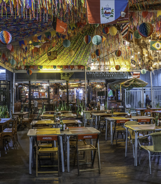 Ein mexikanisches Restaurant mit Tischen, Stühlen und Menschen, das von Hängeschmuck, einem Tor auf der linken Seite und Pflanzen sowie Texttafeln im Hintergrund geprägt ist.