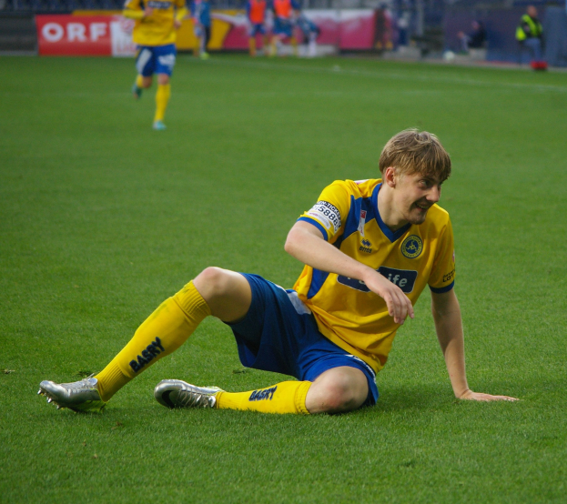 Ein Fußballer sitzt auf dem Boden und hat den Kopf in den Händen, umgeben von Menschen in Sportkleidung, mit unscharfen Hintergrundelementen wie Schildern, Geländern und Pfählen.