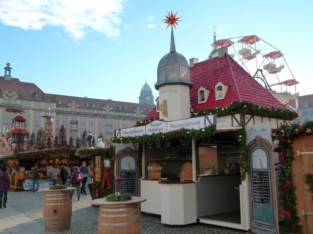 Ein belebter Weihnachtsmarkt in Nürnberg, Deutschland, mit Menschen um dekorierte Stände, festliche Lichter, Schmuck, Gebäude mit Fenstern, ein Riesenrad und einen bewölkten Himmel, mit einer Tafel mit Text auf der rechten Seite.