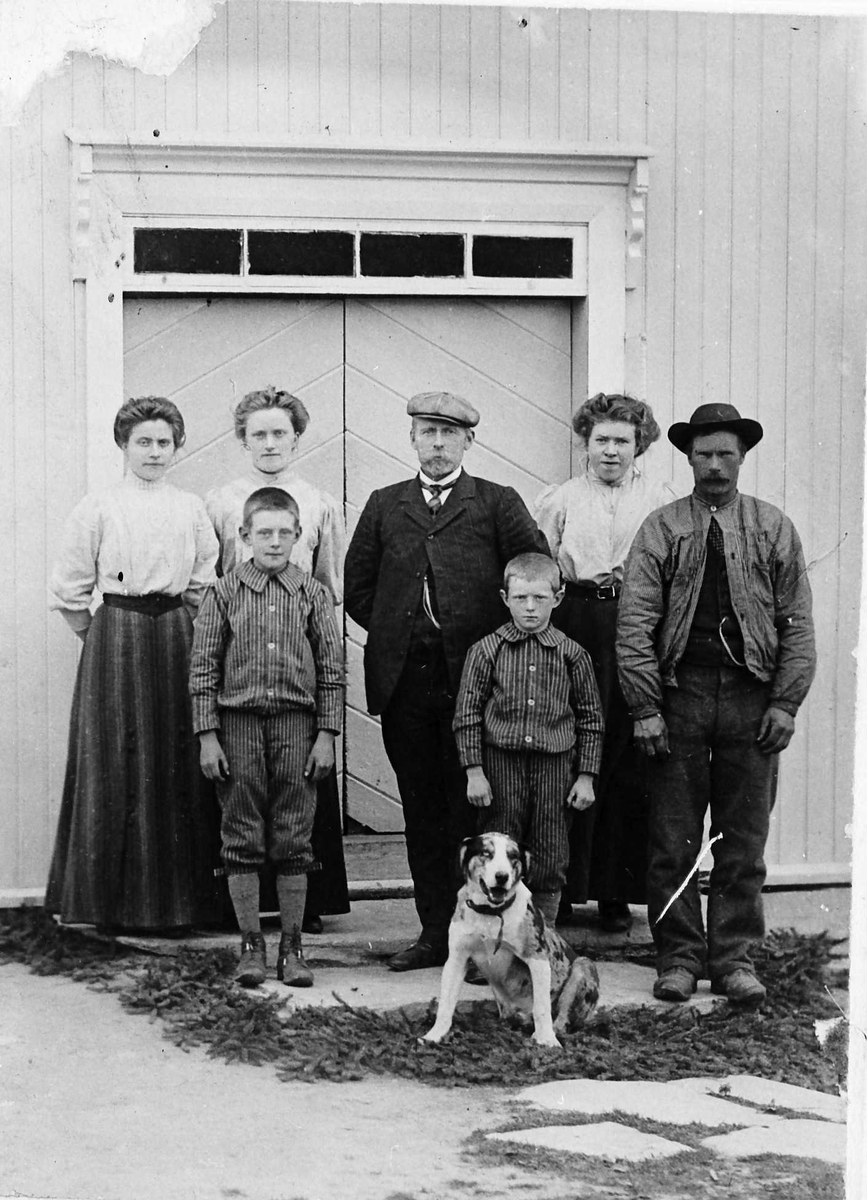 Schwarz-weißes Foto einer Familie mit einem Hund, die vor einem Haus posieren, bestehend aus einem erwachsenen Mann, einer erwachsenen Frau und mehreren Kindern.