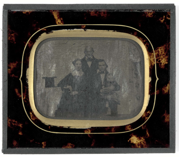 Schwarz-weißes Foto eines Mannes in Anzug und Krawatte und einer Frau in einem unbekannten Outfit, die vor einem alten Fernseher stehen und ernst schauen.