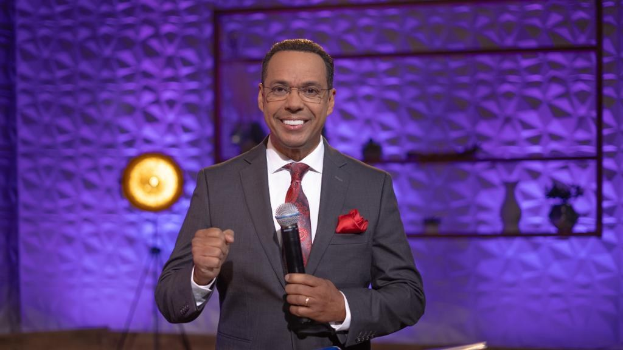 Stephen Colbert, Moderator von 'The Daily Show with Trevor Noah', steht auf einer Bühne und hält ein Mikrofon in der Hand, trägt einen Anzug, Krawatte und eine Brille, während im Hintergrund ein Scheinwerfer und ein Ständer zu sehen sind.