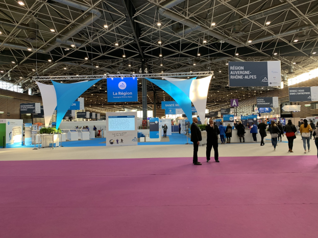 Großer Indoor-Messestand mit zahlreichen Besuchern, einige mit Taschen, unter Deckenbannern, beleuchteten Tischen mit Gegenständen und Deckenbeleuchtung.