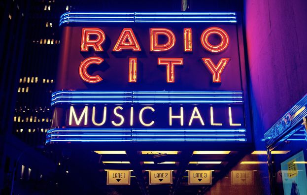 Außenansicht des Radio City Music Hall in New York City bei Nacht mit heller Beleuchtung und hohen umliegenden Gebäuden, mit einer 'Radio City'-Leuchtschrift und zusätzlichen Schildern auf der rechten Seite.