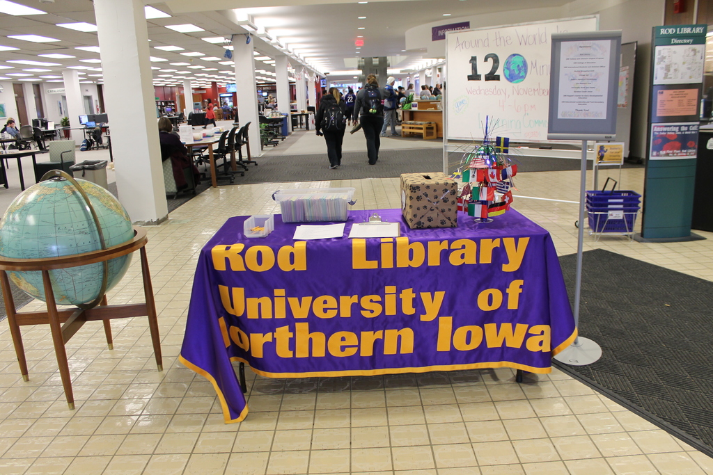 Eine Ausstellung mit einem Tisch, auf dem verschiedene Landesflaggen, eine Kiste, Papiere und ein Banner liegen, eine Kugel zur Linken, ein Anschlagbrett zur Rechten mit einigen Leuten in der Nähe und mehreren Tischen und Stühlen im Hintergrund.