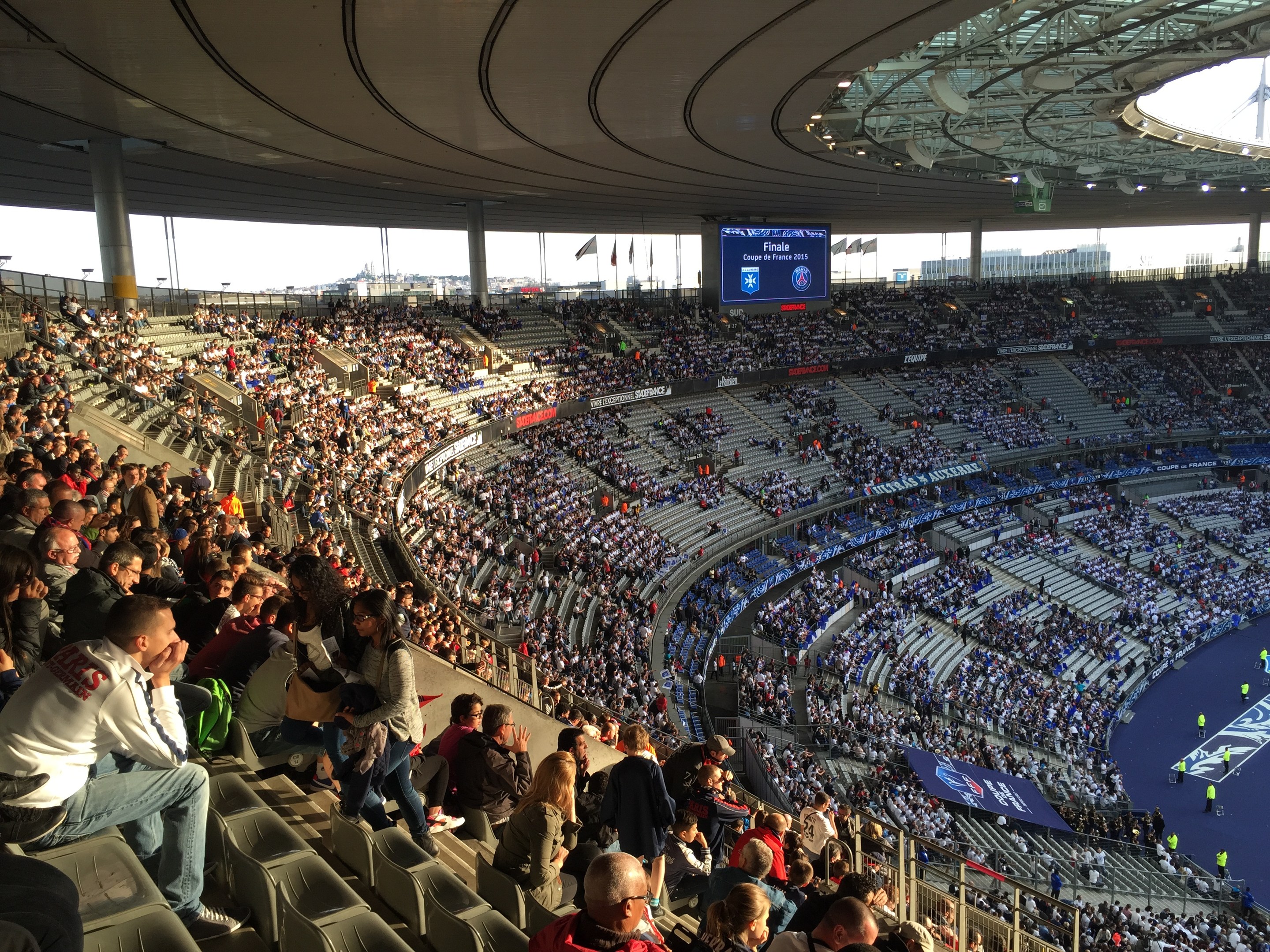 Großes Publikum in einem Stadion bei einem Fötballspiel mit einer Bühne, Fahnen, Masten, einem Bildschirm und der Allianz Arena in München, Deutschland im Hintergrund.