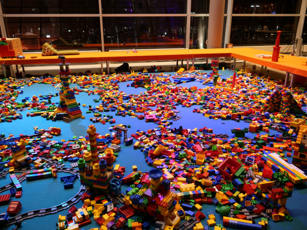 Ein großer Raum mit farbigen LEGO-Bausteinen, die auf dem Boden verstreut sind, umgeben von Tischen und Säulen, durch Glasfenster im Hintergrund sind Menschen und Lichter zu sehen.