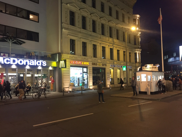 Eine Gruppe von Menschen geht nachts eine Straße in Berlin, Deutschland, entlang mit Gebäuden, Laternen, einer Flagge, Schaufenstern, Fahrrädern und dem Himmel.