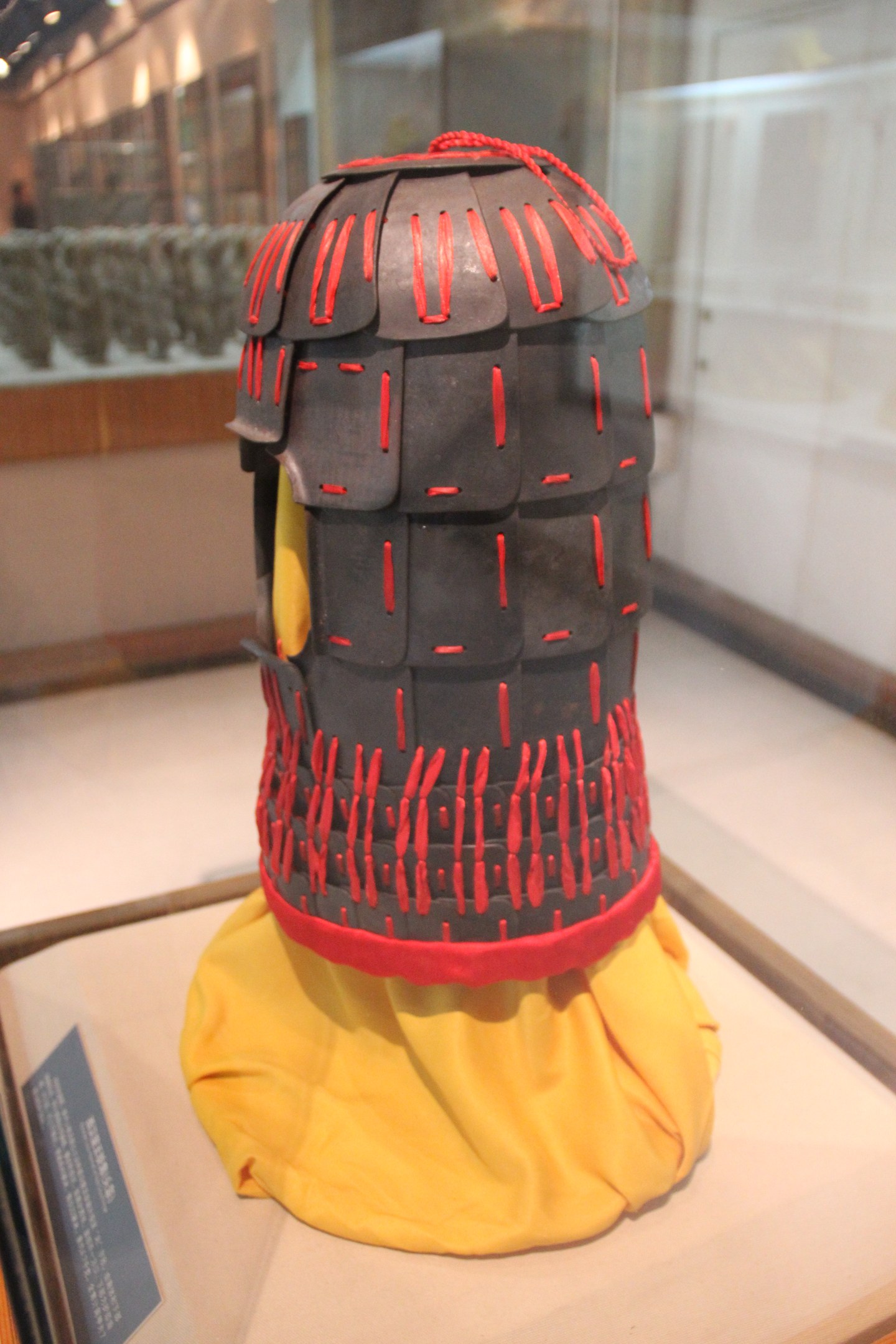 Helm in einer Glasvitrine mit einem gelben Tuch darauf, begleitet von einer Texttafel auf der linken Seite und Deckenlampen im Hintergrund.