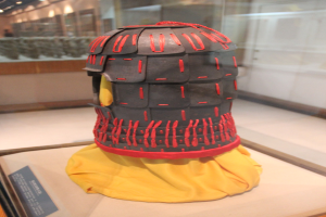 Helm in einer Glasvitrine mit einem gelben Tuch darauf, begleitet von einer Texttafel auf der linken Seite und Deckenlampen im Hintergrund.