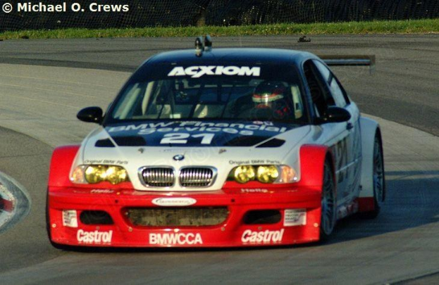 Eine Person in einem Helm fährt ein BMW M3 GT3 Rennauto auf einer von Gras und einem Maschendrahtzaun umgebenen Strecke.