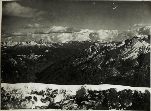 Ein Schwarz-Weiß-Foto einer Gebirgskette mit schneebedeckten Gipfeln, Bäumen und Wolken am Himmel, mit Text unten lesend "Gustav Klimt - Die Alpen, 1902".