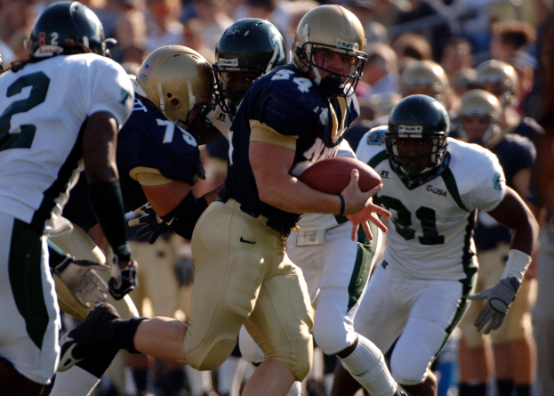 A football player in motion with the ball, surrounded by opponents and teammates in sports attire and helmets, with a blurred background indicating speed.