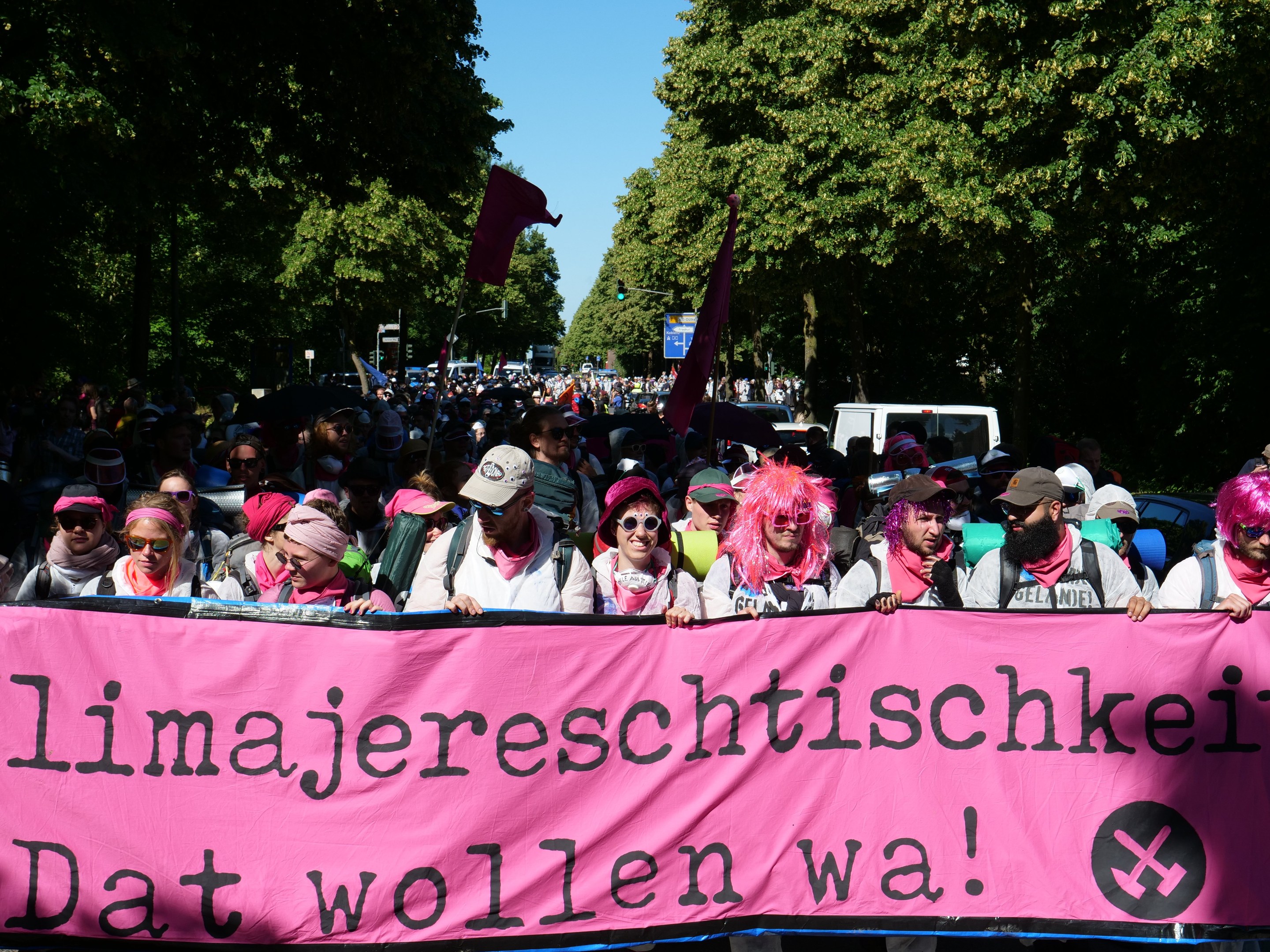 Eine Gruppe von Menschen marschiert auf einer Straße, hält ein pinkes Banner mit der Aufschrift "Women's March in Berlin", wobei einige eine Mütze und eine Schutzbrille tragen und andere Fahnen halten, vor einem Hintergrund aus Bäumen, Fahrzeugen und Himmel.