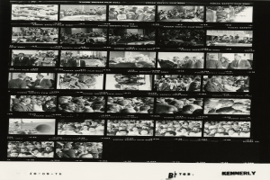 Schwarzes und weißes Foto einer vielseitigen Menge strahlender Menschen, die um einen Fernseher versammelt sind und in die Kamera schauen.