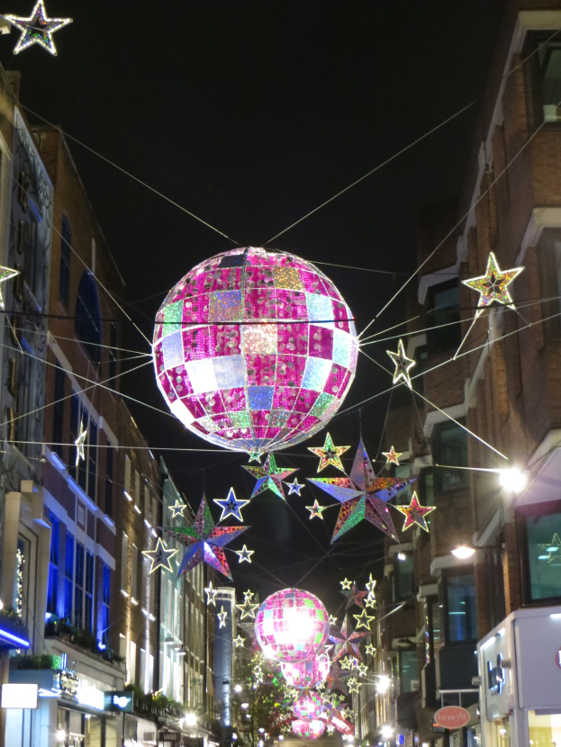 Eine festlich geschmückte Stadtstraße bei Nacht mit einem großen Diskokugel an einem Gebäude hängend und beleuchteten Dekorationen auf den umliegenden Gebäuden.