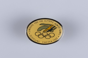 Ein runder Abzeichenaufkleber mit schwarzem Hintergrund und goldenem Olympische Winterspiele-Logo, mittig auf einer weißen Oberfläche platziert, mit fetter weißer Schrift, die "Olympische Winterspiele" lautet.