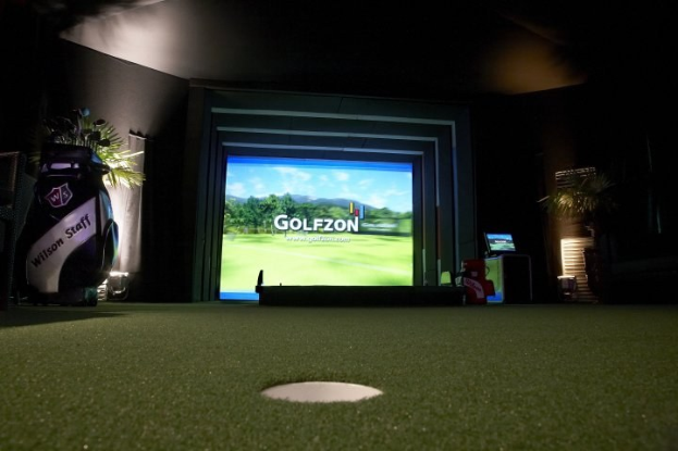 Golfsimulator mit großem Bildschirm, der eine Golfanlage zeigt, flankiert von einer Golftasche und einer Pflanze auf der linken Seite und anderen Gegenständen auf der rechten Seite, vor dunklem Hintergrund.