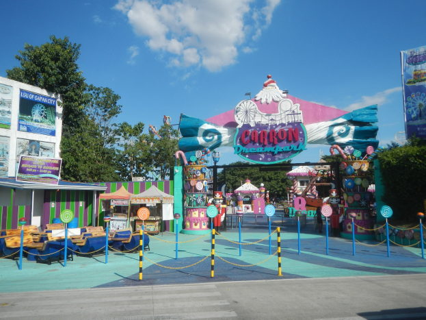 Kirmesfahrgeschäft am Parkeingang mit Straße, Umzäunung, Buden, Banner, Bäumen und bewölktem Himmel im Hintergrund.