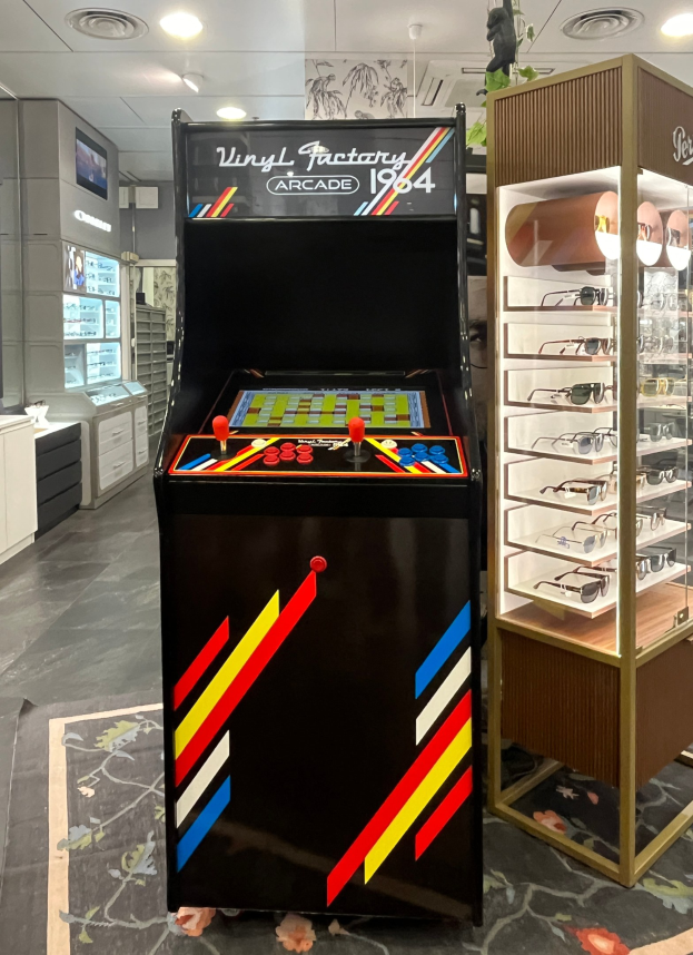 Ein Geschäft mit einem zentralen Vintage-Arcade-Automaten, einem rechten Glasvitrine und Hintergrund-Elementen wie Schränken, einem Fernseher und anderen Gegenständen, mit Deckenlampen, die eine Retro-Atmosphäre schaffen.