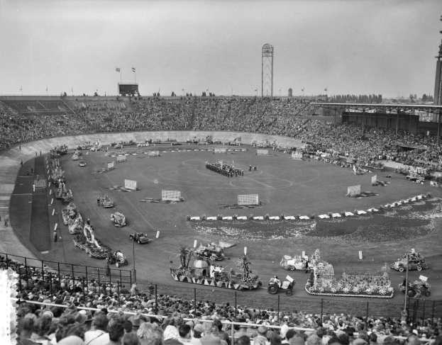 Schwarzes und weißes Bild einer großen Menschenmenge, die ein Rennen in einem Stadion mit Fahrzeugen, Zuschauern, Absperrungen, Pfählen, Fahnen, Türmen, Bäumen und Himmel sieht.