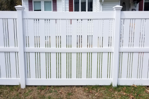 Eine weiße Vinyl-Privatsichtschutzzaun steht vor einem Haus mit Fenstern, umgeben von Gras und trockenen Blättern, mit Bäumen im Hintergrund.