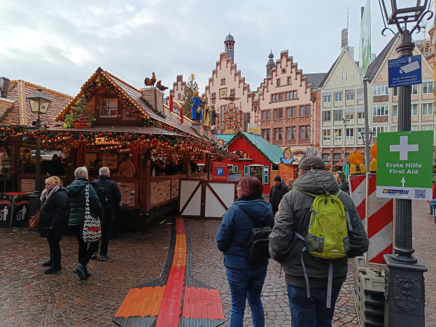 Eine Gruppe von Menschen, die auf einem Kopfsteinpflasterweg neben einem Weihnachtsmarkt in Nürnberg, Deutschland, entlanggehen, mit Laternenmasten, Texttafeln und Gebäuden im Hintergrund unter einem bewölkten Himmel.