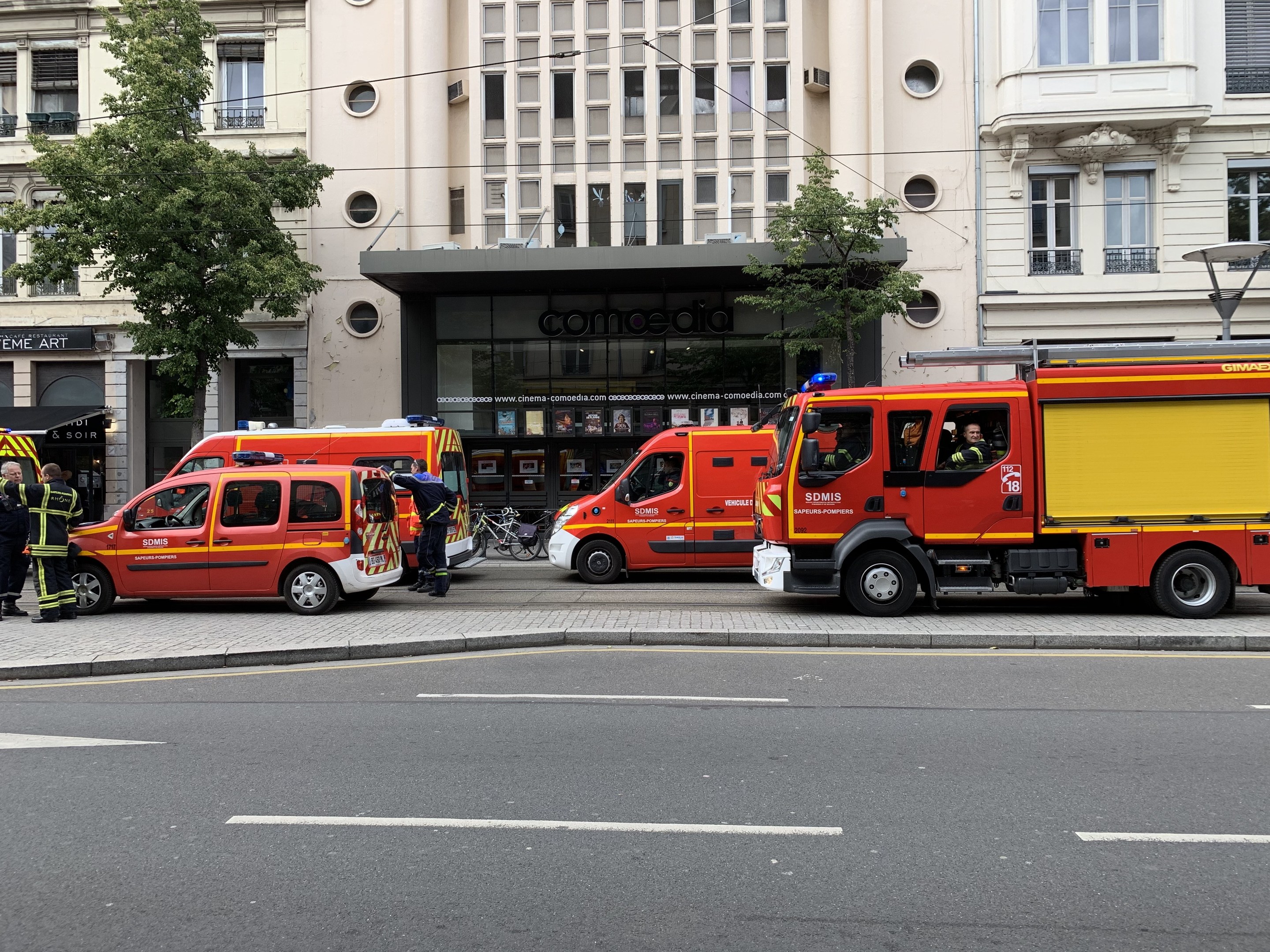 Eine Gruppe von Feuerwehrautos auf einer Pariser Straße geparkt mit Menschen in der Nähe, Gebäuden, Bäumen und einem Fahrrad im Hintergrund, wahrscheinlich an einem Unfallort.