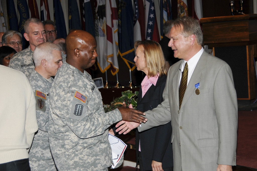 Eine Gruppe von Menschen, die mit zwei Personen Handsch├╝tteln, Fahnen im Hintergrund, ein Podium mit Mikrofon und eine Topfpflanze in der N├Ąhe.