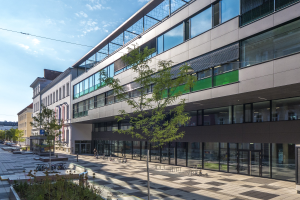 Großes modernes Gebäude mit vielen Fenstern, umgeben von Bäumen, Pflanzen, Pfählen, Fahrzeugen und Menschen unter einem bewölkten Himmel, identifiziert als die neue Hauptquartier des Deutschen Instituts für Technologie in Berlin.