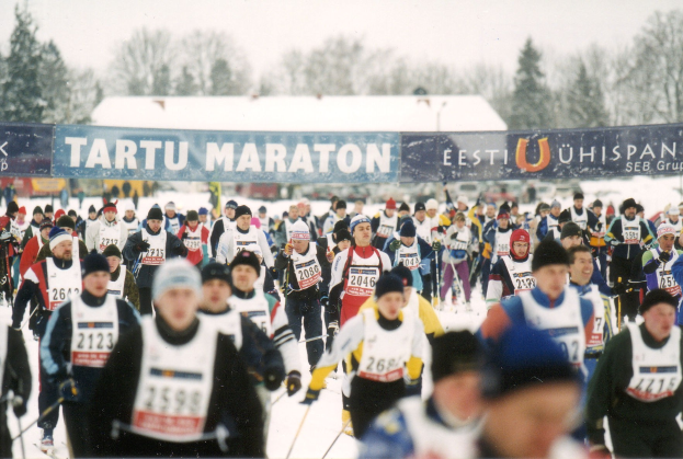 Gruppe von Menschen, die auf Skiern rennt und Mützen trägt, mit Ski-Stöcken, Bannern, Bäumen und einem klaren blauen Himmel im Hintergrund.
