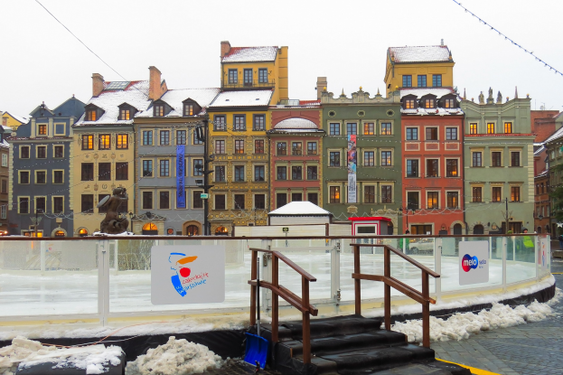 Eisbahn in einem Stadtplatz umgeben von Gebäuden, Treppen, Geländern, Informationsschildern, schneebedecktem Boden, Laternen, Bäumen und einem Himmel als Hintergrund.