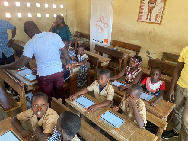 Kinder sitzen an Tischen in einem Klassenzimmer und nutzen Tablets, während Erwachsene in der Nähe stehen und Bildungsplakate und ein Banner an den Wänden zu sehen sind.
