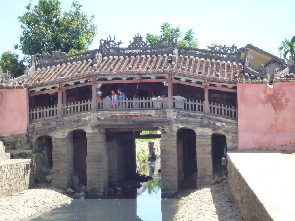 Menschen auf einer Brücke mit einem Zaun und Skulpturen, Bäume im Hintergrund und Wasser darunter.