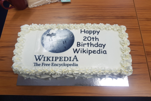Ein Geburtstagskuchen mit "Herzlichen Glückwunsch zum 20. Geburtstag Wikipedia" darauf, der auf einem Tisch mit einer Tasse, einer Abdeckung und anderen Gegenständen steht.