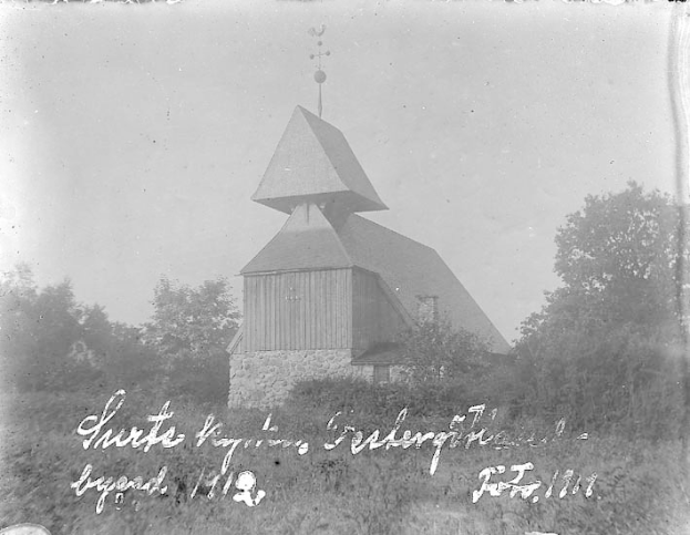Schwarzes und weißes Foto einer Kirche mit einem Turm, umgeben von Bäumen und Pflanzen, unter einem bewölkten Himmel, beschriftet mit "Kirche des Heiligen Grabes" unten.