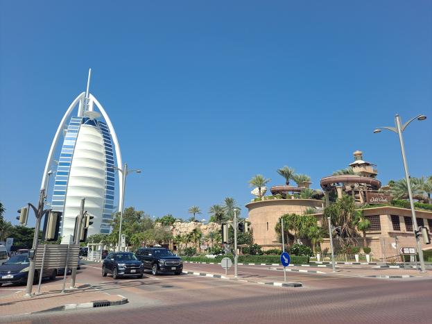 Das Burj Al Arab Hotel in Dubai, VAE, mit Fahrzeugen, Straßeninfrastruktur und Gebäuden im Vordergrund vor einem Himmelhintergrund.
