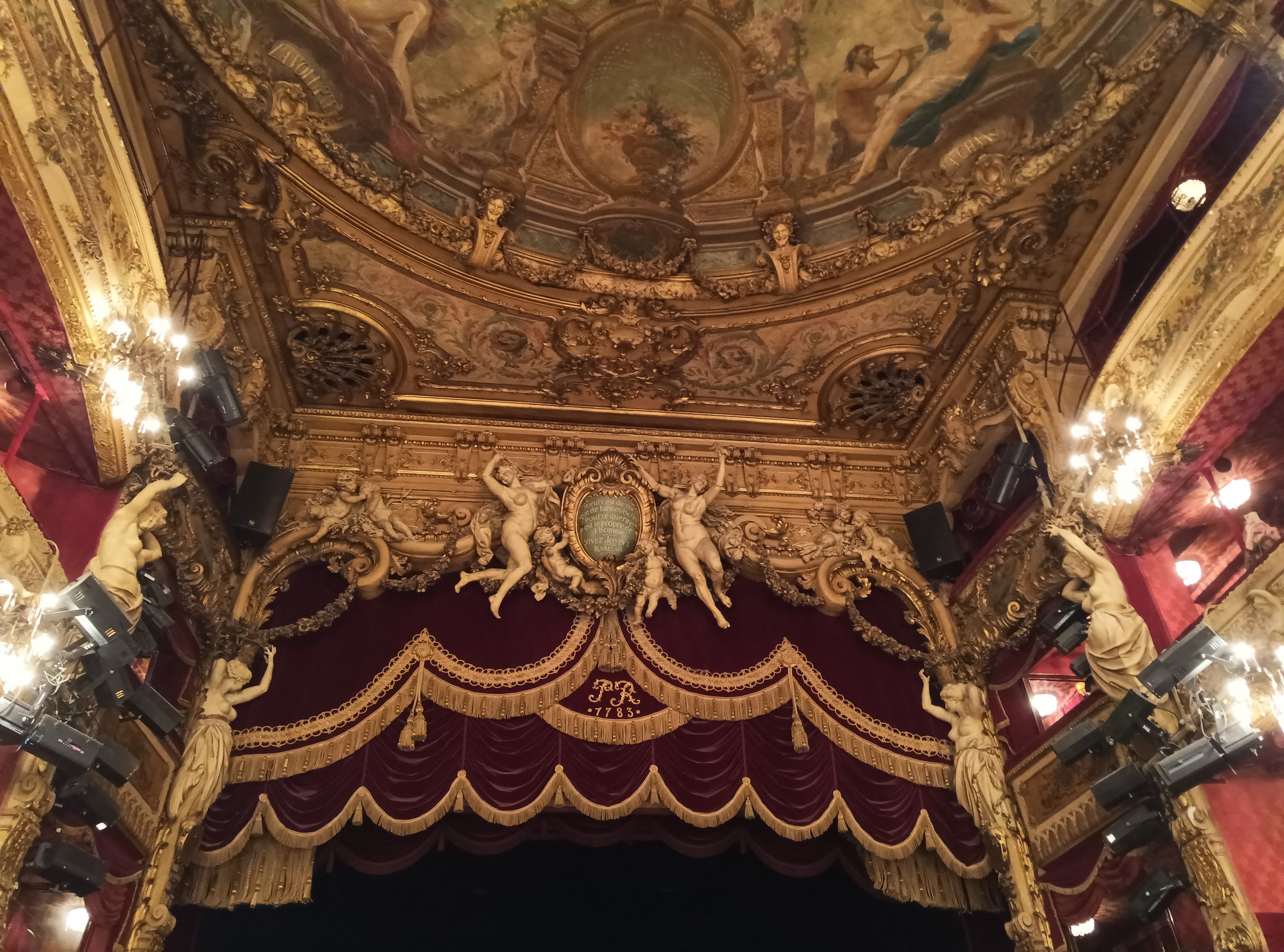 Das Innere eines Theaters mit rotem Vorhang, Kronleuchtern, Skulpturen, Lichtern und einer Deckenmalerei, das eine Bühne mit ein paar Menschen unten zeigt.