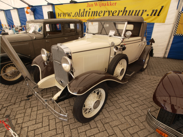 Ein vintage-Rummelplatz-Auto, das vor einem blauen und weißen Zelt mit einer Fahne und Luftballons im Hintergrund geparkt ist.