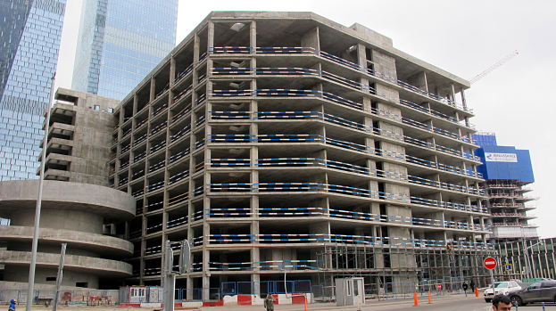 Großer Bau in einer städtischen Umgebung mit Wolkenkratzern, Fahrzeugen, Fußgängern, Baumaschinen und Beschilderung vor einem klaren Himmel.
